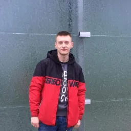 Młody mężczyzna w kurtce w kolorach czerwonym i czarnym, stojący na tle ściany z zielonych paneli ze szklanymi drzwiami.