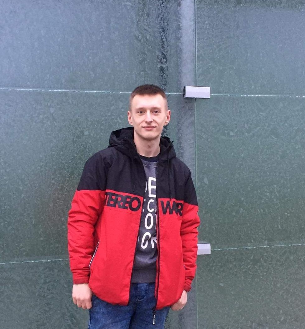 Młody mężczyzna w kurtce w kolorach czerwonym i czarnym, stojący na tle ściany z zielonych paneli ze szklanymi drzwiami.