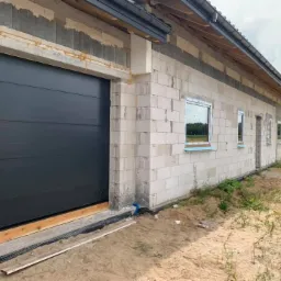 Nowoczesna, grafitowa brama segmentowa w surowym stanie budynku z białych bloczków, widoczne okna z niebieską folią ochronną i niezabezpieczone krawędzie muru.