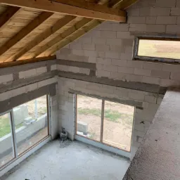 Surowy stan domu w budowie: widok z góry na wstawione okna w ścianach z białych bloczków, drewniana konstrukcja dachu, widoczny krajobraz za oknami.