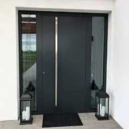 Nowoczesne, grafitowe drzwi wejściowe z bocznymi przeszkleniami, długim, pionowym uchwytem ze stali nierdzewnej, oświetlone lampionami ze świecami, na jasnej kostce brukowej.