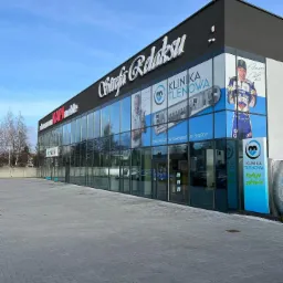 Duży, nowoczesny budynek z przeszkloną fasadą, z widocznymi reklamami na oknach, w tym nazwa 'Strefa Relaksu' i 'Klinika Tlenowa', z zewnętrznymi drzwiami wejściowymi i szarym brukowanym chodnikiem.