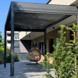 Nowoczesna pergola z regulowanymi lamelami w kolorze antracytowym, z wiszącym fotelem kokonem i zadbaną zielenią w tle, na tarasie z szarych płytek.