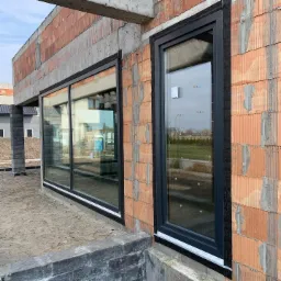 Montaż czarnych ram okiennych w surowym budynku z czerwonej cegły, widoczne zabezpieczenia montażowe i fragmenty budynku w trakcie budowy.