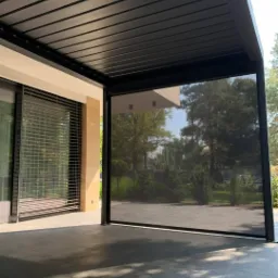 Zadaszony taras z czarną konstrukcją, wyposażony w roletę zewnętrzną z tkaniny zaciemniającej, widok na ogród.