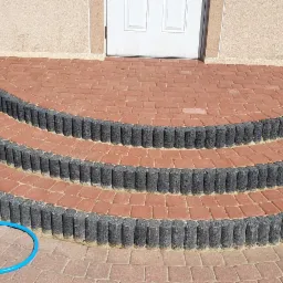 Półokrągłe schody wejściowe wyłożone czerwoną kostką brukową, obrzeża z szarych, betonowych palisad. W tle białe drzwi i beżowa elewacja budynku. Z boku widoczny zwój niebieskiego węża ogrodowego...