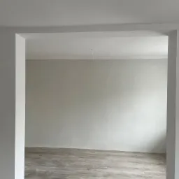 Wnętrze po malowaniu na szaro, widoczne drewnopodobne panele podłogowe, białe ściany i sufit z listwą dekoracyjną, wystający kabel oświetleniowy.