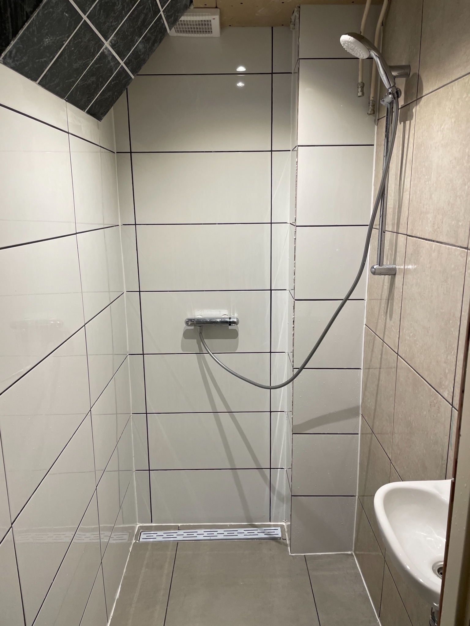 Wykończona kabina prysznicowa z białymi płytkami i czarnymi fugami, z odpływem liniowym i baterią termostatyczną, z widoczną umywalką po prawej stronie i skośnym sufitem wyłożonym czarnymi płytkami.