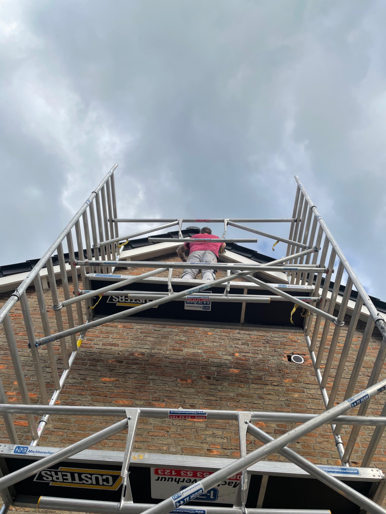 Pracownik na rusztowaniu przy murowanej elewacji, widok z dołu na górę, pochmurne niebo w tle.