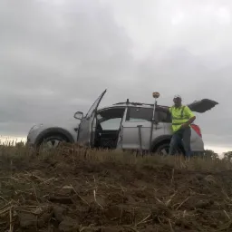 Srebrny SUV z otwartymi drzwiami zaparkowany na zaoranym polu, obok mężczyzna w żółtej kamizelce i białej czapce opierający się o auto z zamontowanym na dachu sprzętem pomiarowym.