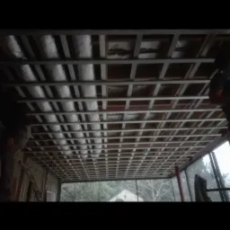 Montaż metalowej konstrukcji sufitu podwieszanego z izolacją termiczną, widok z dołu na szkielet i częściowo ułożoną wełnę mineralną, okno z widokiem na zewnątrz w tle.