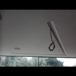 Niedokończony sufit podwieszany z wystającym kablem i otworem na głośnik, widok na drzewa za oknem.