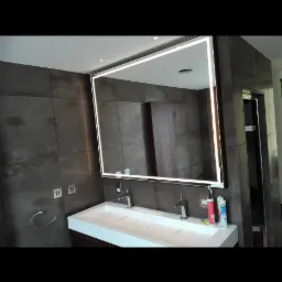 Nowoczesna łazienka z podwójną umywalką, dużym lustrem z podświetleniem LED i szarą płytką imitującą kamień na ścianie. Widoczne akcesoria łazienkowe i dozowniki na blacie.