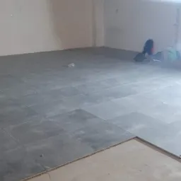 Szare płytki podłogowe ułożone częściowo w pustym, jasnym pomieszczeniu, widoczna warstwa wyrównująca podłoże przed położeniem płytek, torby robocze w rogu.