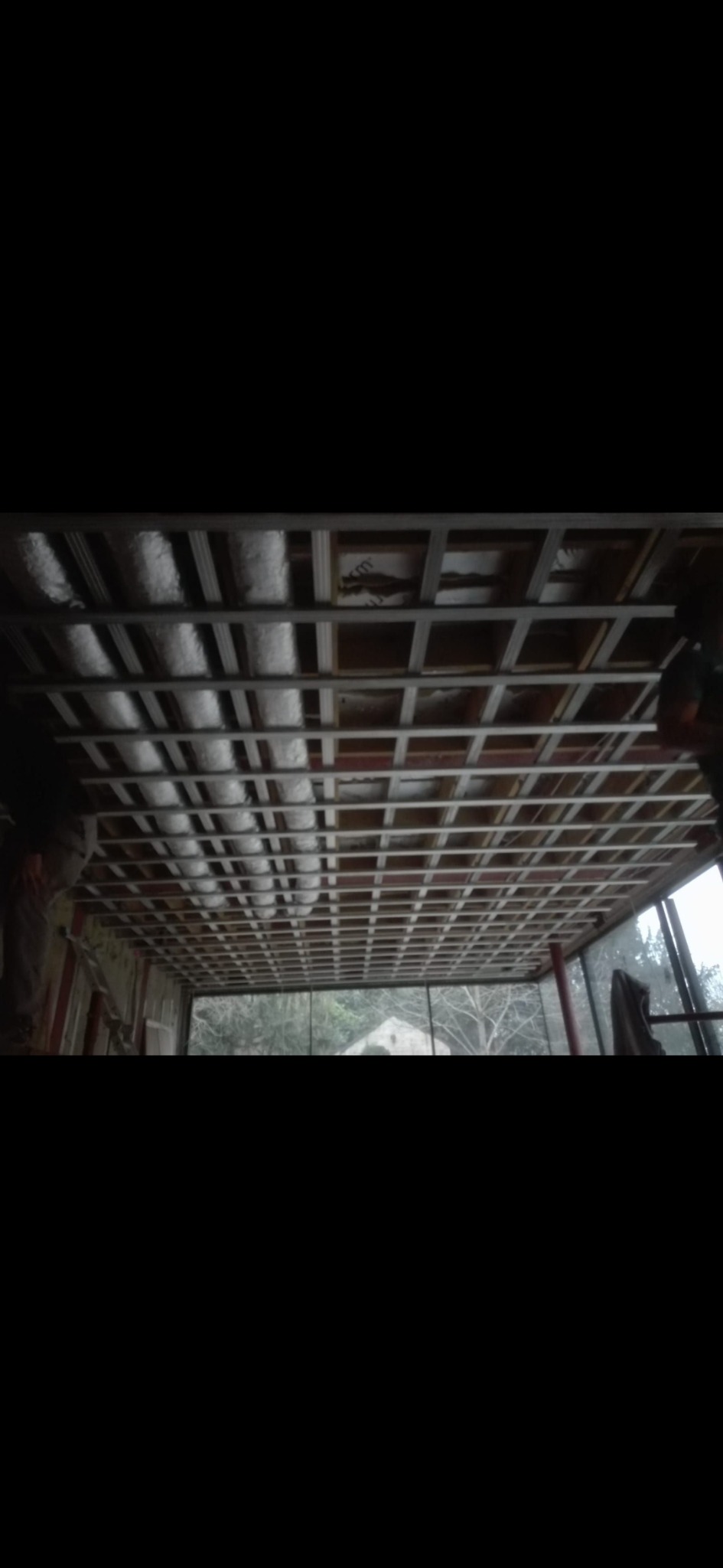 Montaż metalowej konstrukcji sufitu podwieszanego z izolacją termiczną, widok z dołu na szkielet i częściowo ułożoną wełnę mineralną, okno z widokiem na zewnątrz w tle.