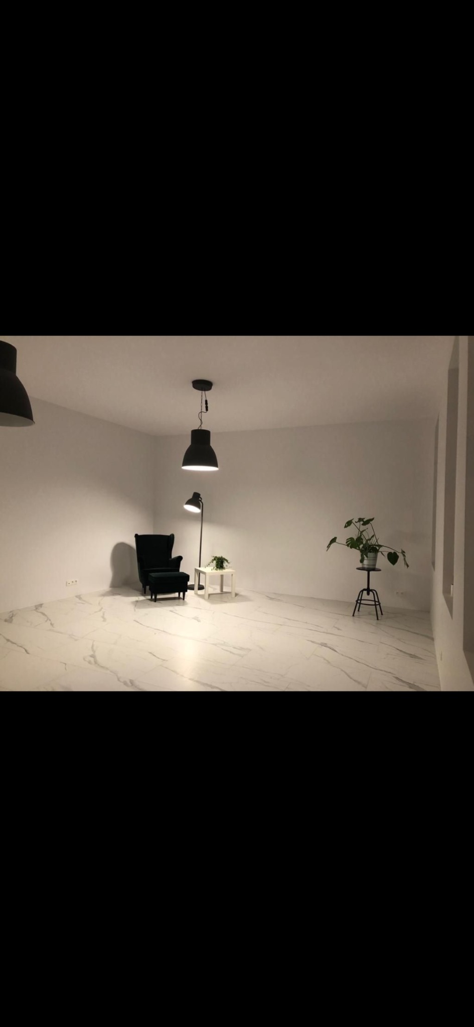 Jasne wnętrze z marmurową podłogą, ciemnozielonym fotelem z podnóżkiem, lampą podłogową i roślinami na stolikach.