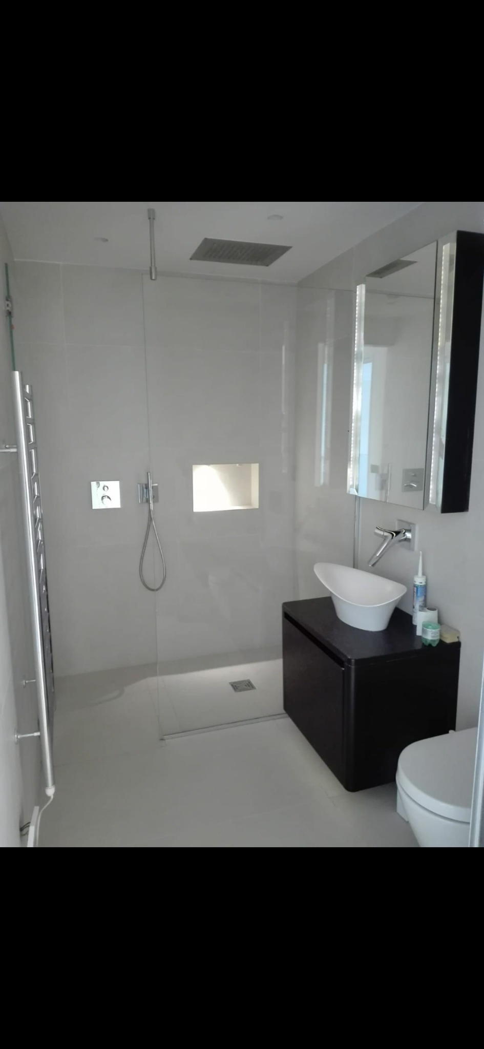 Nowoczesna łazienka z szarymi płytkami, przeszklonym prysznicem typu walk-in, wiszącą szafką z asymetryczną umywalką i grzejnikiem drabinkowym.