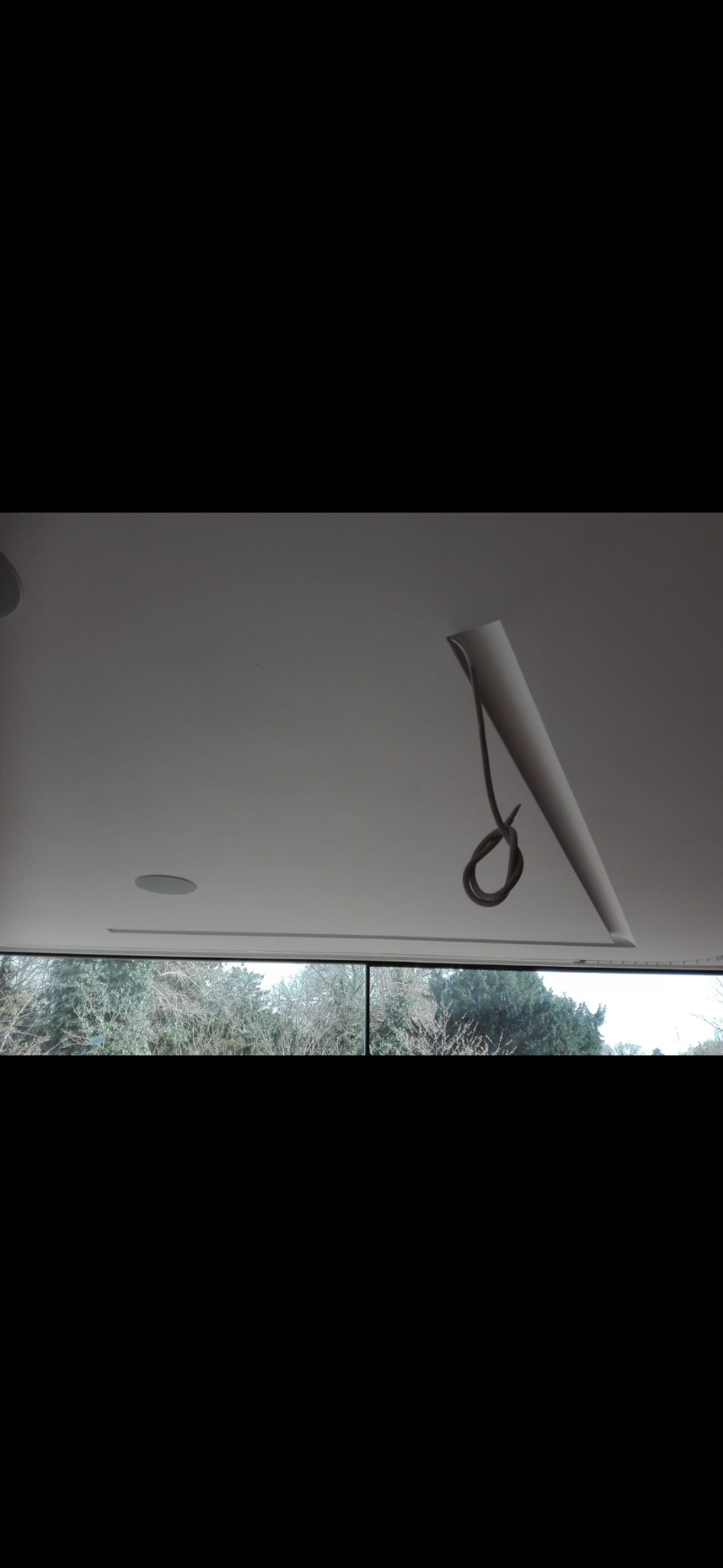 Niedokończony sufit podwieszany z wystającym kablem i otworem na głośnik, widok na drzewa za oknem.