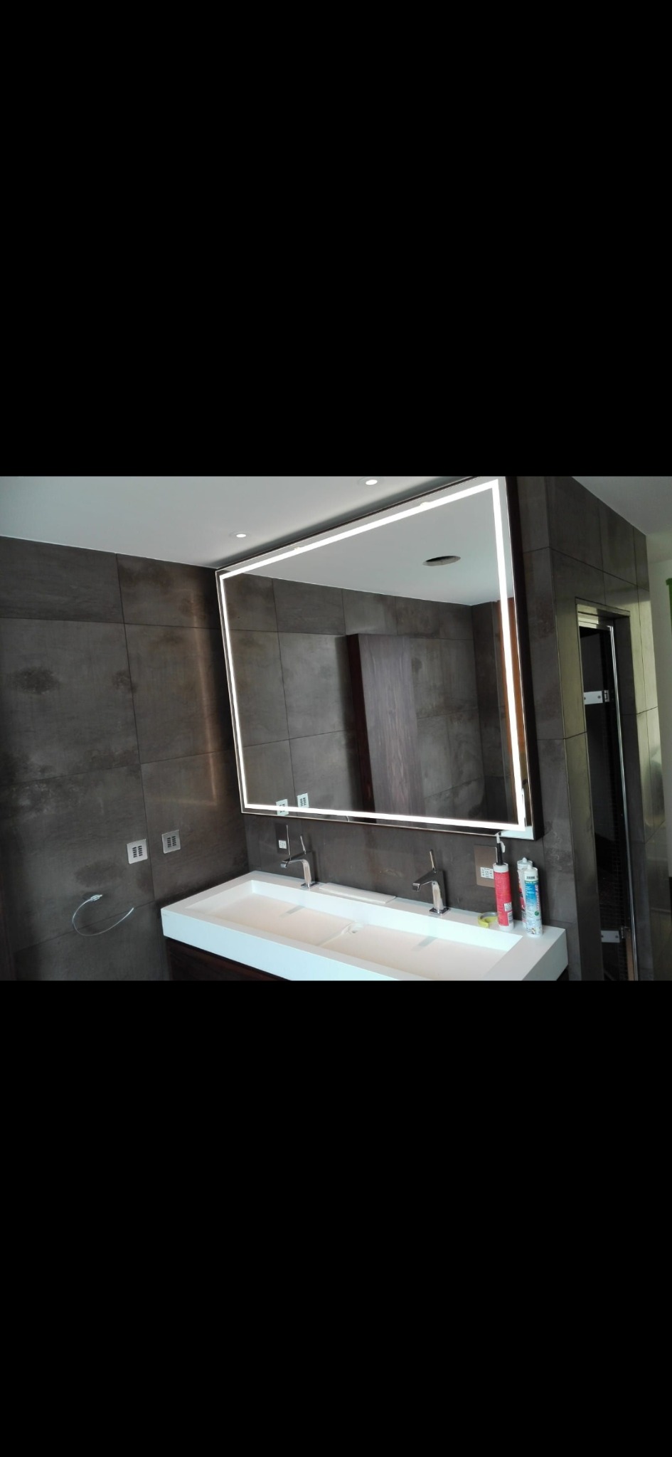 Nowoczesna łazienka z podwójną umywalką, dużym lustrem z podświetleniem LED i szarą płytką imitującą kamień na ścianie. Widoczne akcesoria łazienkowe i dozowniki na blacie.