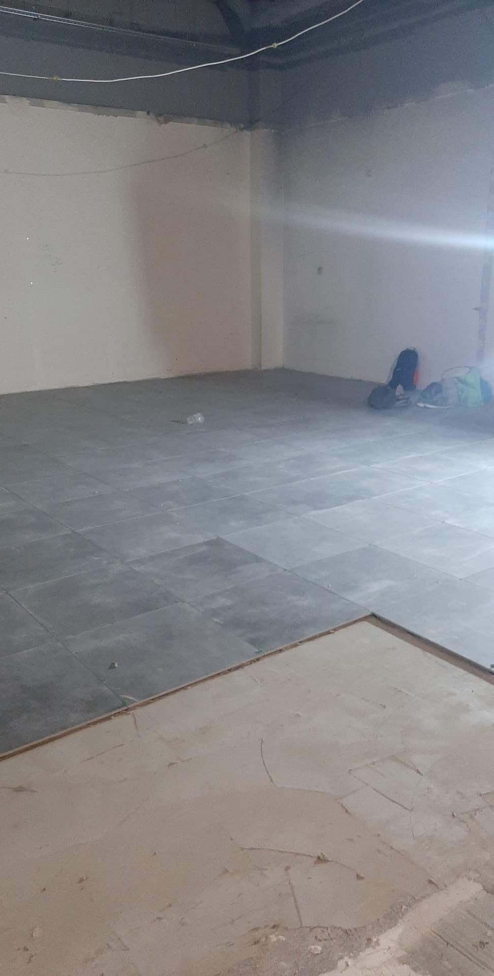 Szare płytki podłogowe ułożone częściowo w pustym, jasnym pomieszczeniu, widoczna warstwa wyrównująca podłoże przed położeniem płytek, torby robocze w rogu.