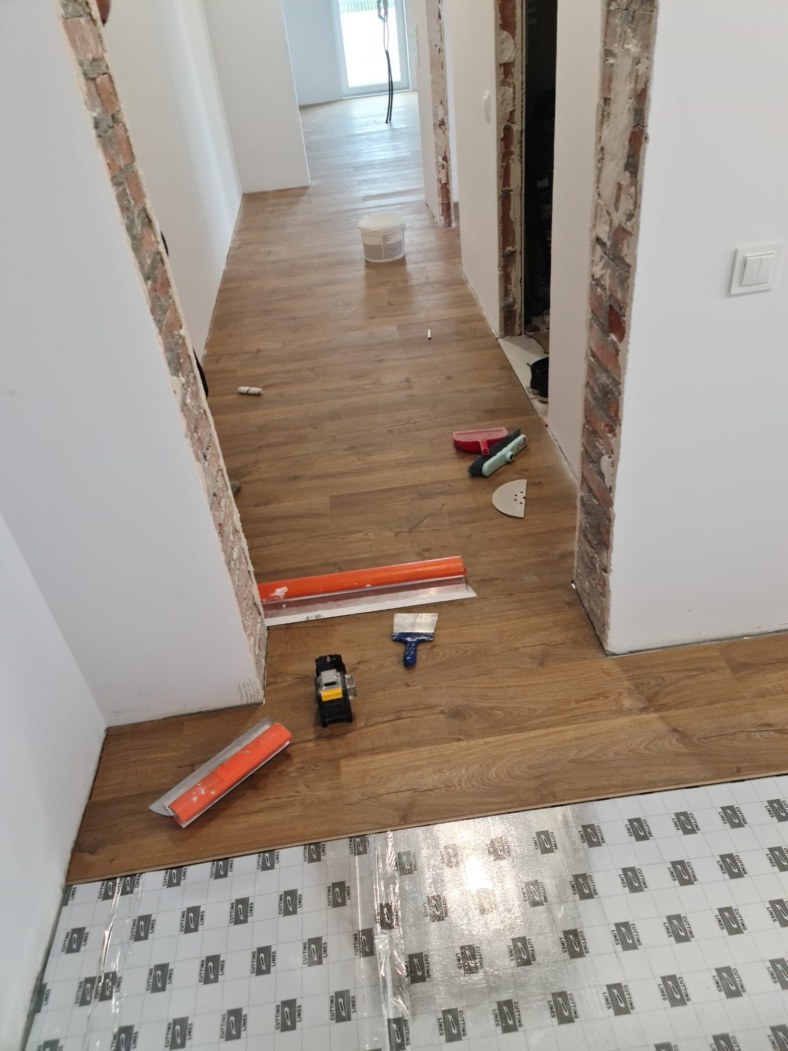 Układanie paneli podłogowych na folii izolacyjnej, widoczne narzędzia glazurnicze i odsłonięta cegła w ościeżnicy drzwi.