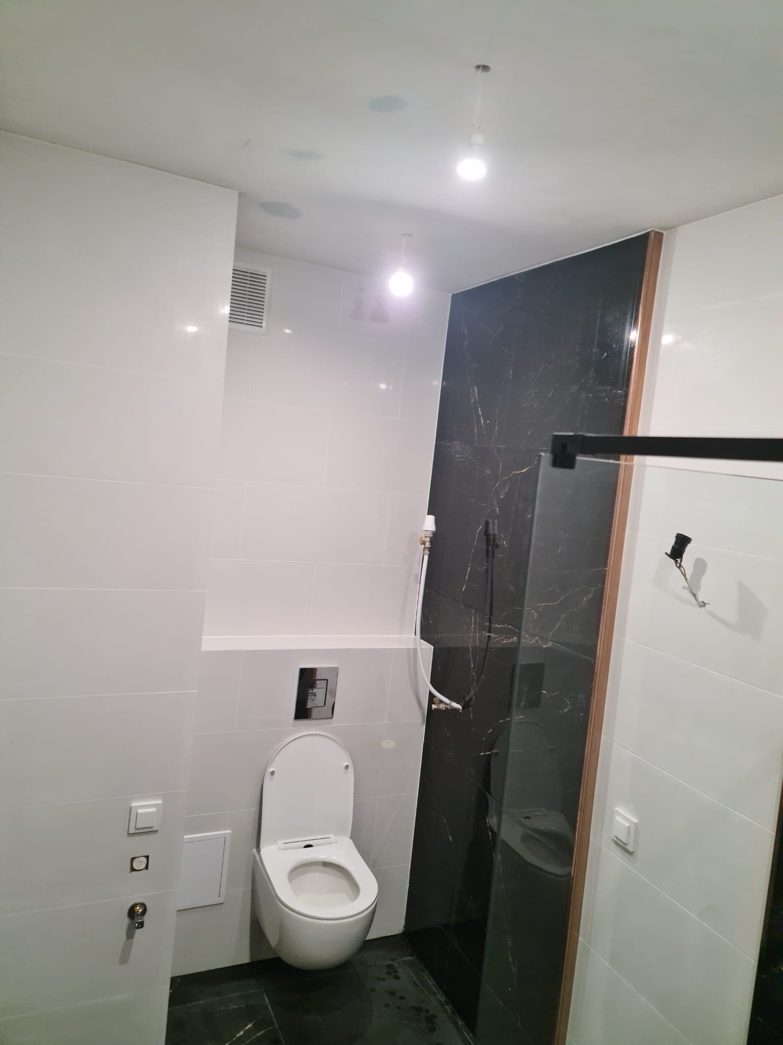 Nowoczesna łazienka z białymi płytkami, toaleta podtynkowa, czarny marmurkowy panel prysznicowy z drewnianą ramą i czarną armaturą, oświetlenie punktowe.