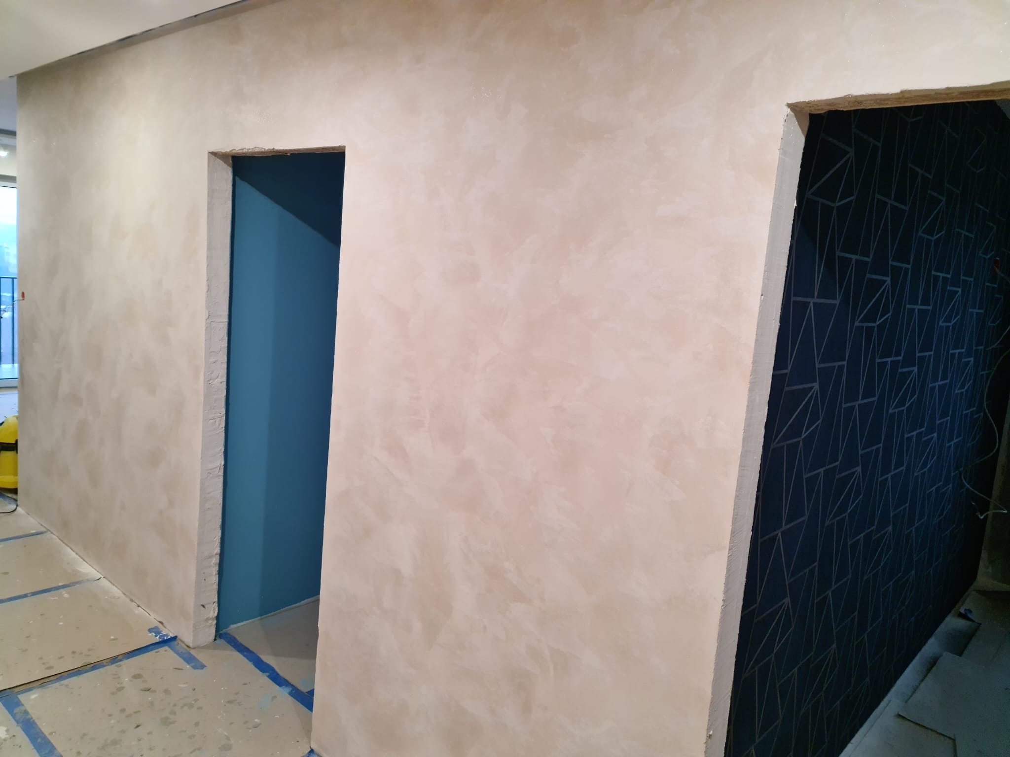 Ściana z efektem dekoracyjnym w odcieniach beżu i dwa otwory drzwiowe: jeden z pomalowanymi na niebiesko ścianami, drugi z tapetą w geometryczne wzory. Podłoga zabezpieczona folią malarską.