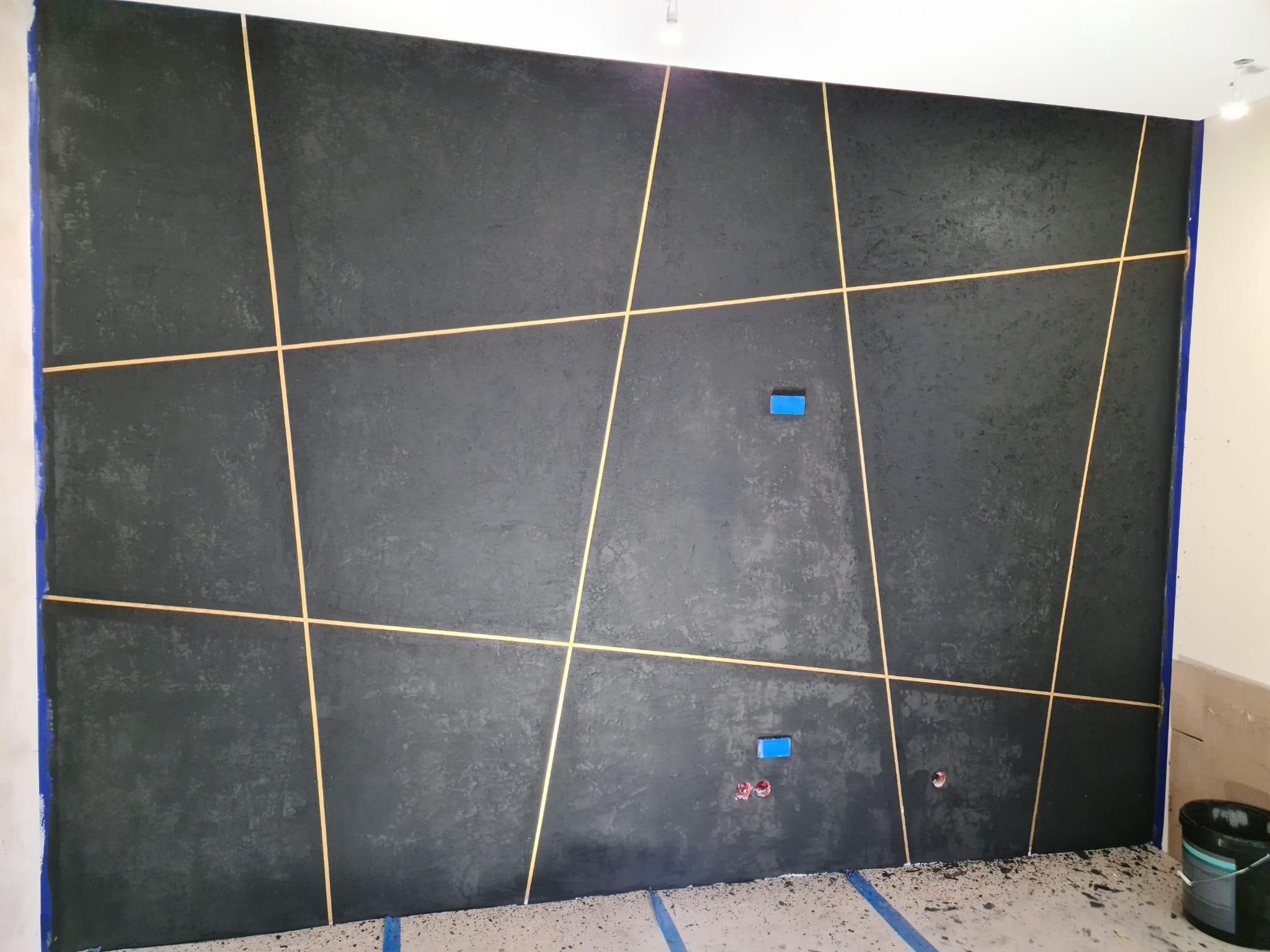 Ściana w ciemnym, grafitowym odcieniu z dekoracyjnymi, złotymi listwami tworzącymi geometryczny wzór. Niebieska taśma malarska zabezpiecza krawędzie. Widoczne gniazdka elektryczne i ślady po...
