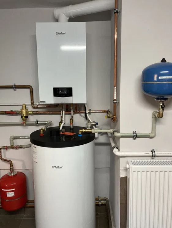 Zestaw grzewczy Vaillant: wiszący piec gazowy, zasobnik ciepłej wody z czarnym blatem, czerwone naczynie przeponowe i niebieski zbiornik wyrównawczy, połączone rurami miedzianymi i plastikowymi...