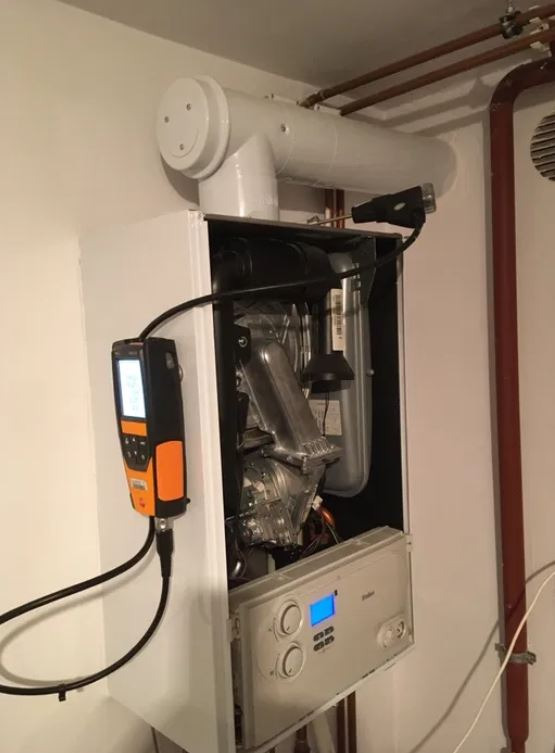 Biały, wiszący piec gazowy z otwartą obudową, podłączony do analizatora spalin z pomarańczowymi elementami, podczas pomiaru parametrów spalania.