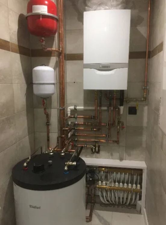 Kompletna instalacja centralnego ogrzewania z kotłem gazowym, zasobnikiem ciepłej wody i rozdzielaczem podłogówki, wykonana z miedzianych rur na tle beżowych płytek.