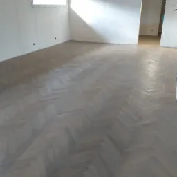 Wykończony parkiet ułożony w jodełkę w jasnym, pustym pokoju z białymi ścianami i otwartymi drzwiami do kolejnego pomieszczenia.