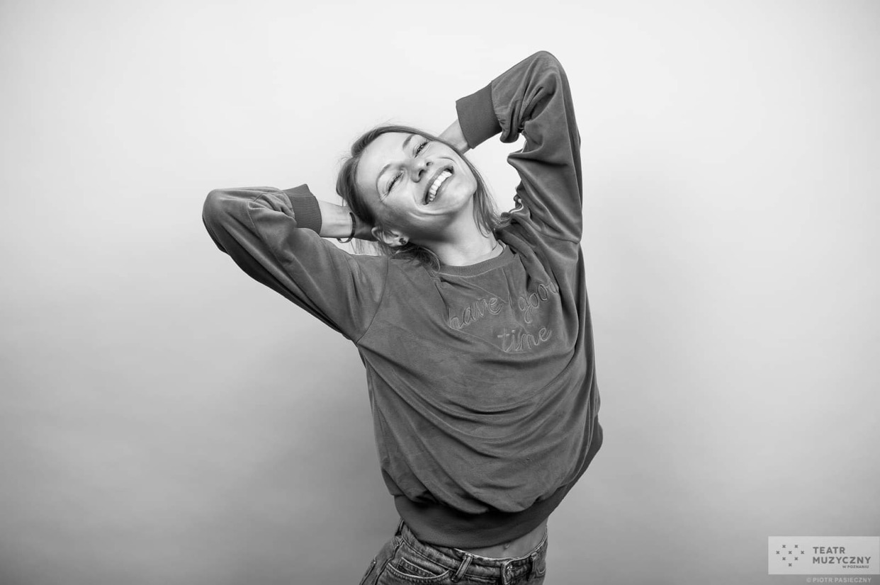 Czarno-białe zdjęcie uśmiechniętej kobiety z rękami założonymi za głowę, ubrana w bluzę z napisem 'have good time', pochylona do tyłu.