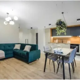 Wnętrze otwartej przestrzeni: szmaragdowa sofa narożna z beżowym pledem, stół z czterema szarymi krzesłami, zabudowa kuchenna z wiszącą rośliną doniczkową i drewnianym panelem nad blatem, parkiet.