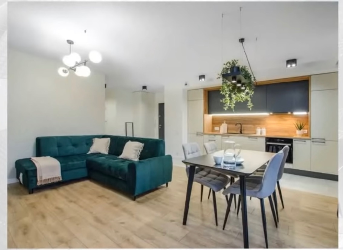 Wnętrze otwartej przestrzeni: szmaragdowa sofa narożna z beżowym pledem, stół z czterema szarymi krzesłami, zabudowa kuchenna z wiszącą rośliną doniczkową i drewnianym panelem nad blatem, parkiet.