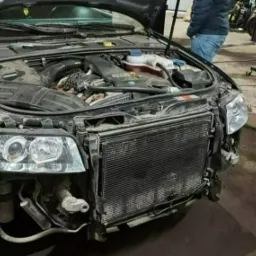 Szary samochód z otwartą maską na podnośniku w warsztacie, widoczny silnik i chłodnica, zdemontowane elementy karoserii.