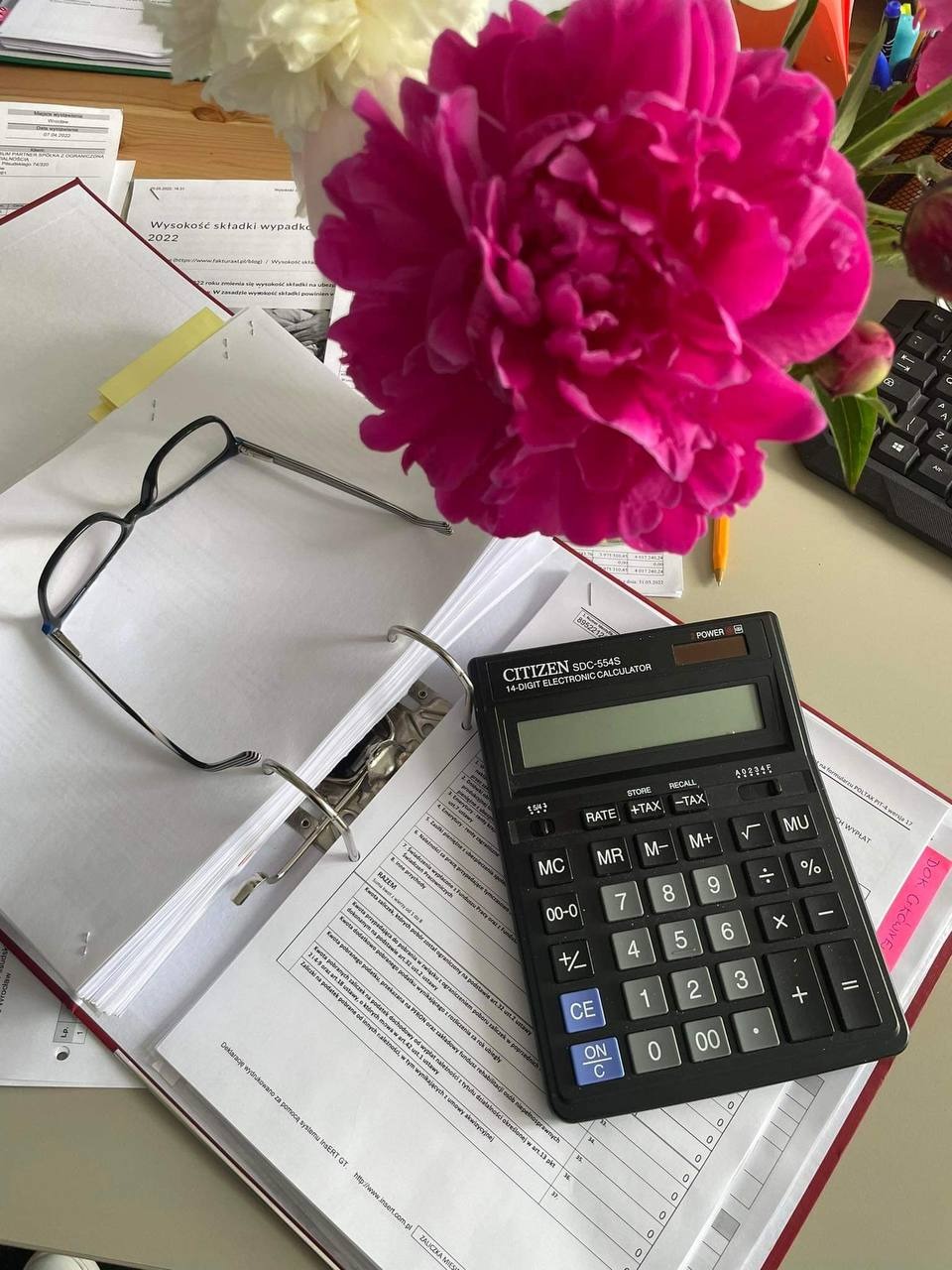Kalkulator Citizen SDC-554S, okulary i otwarty segregator z dokumentami na biurku, obok wazon z piwoniami, tworzące scenerię pracy księgowej.