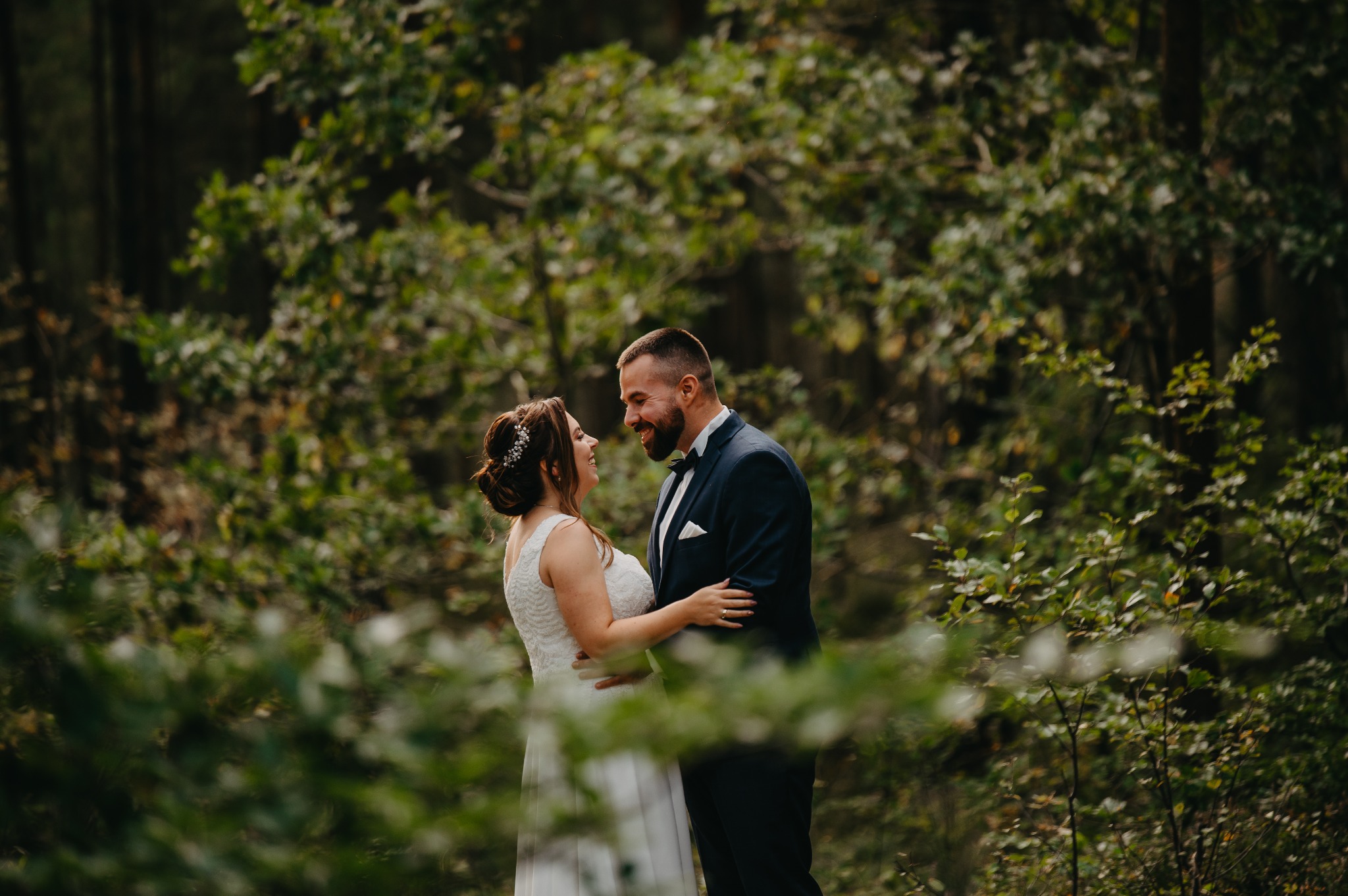 Uśmiechnięta para młoda w objęciach na tle zieleni leśnej, panna młoda w koronkowej sukni z upiętymi włosami, pan młody w granatowym garniturze z muchą.