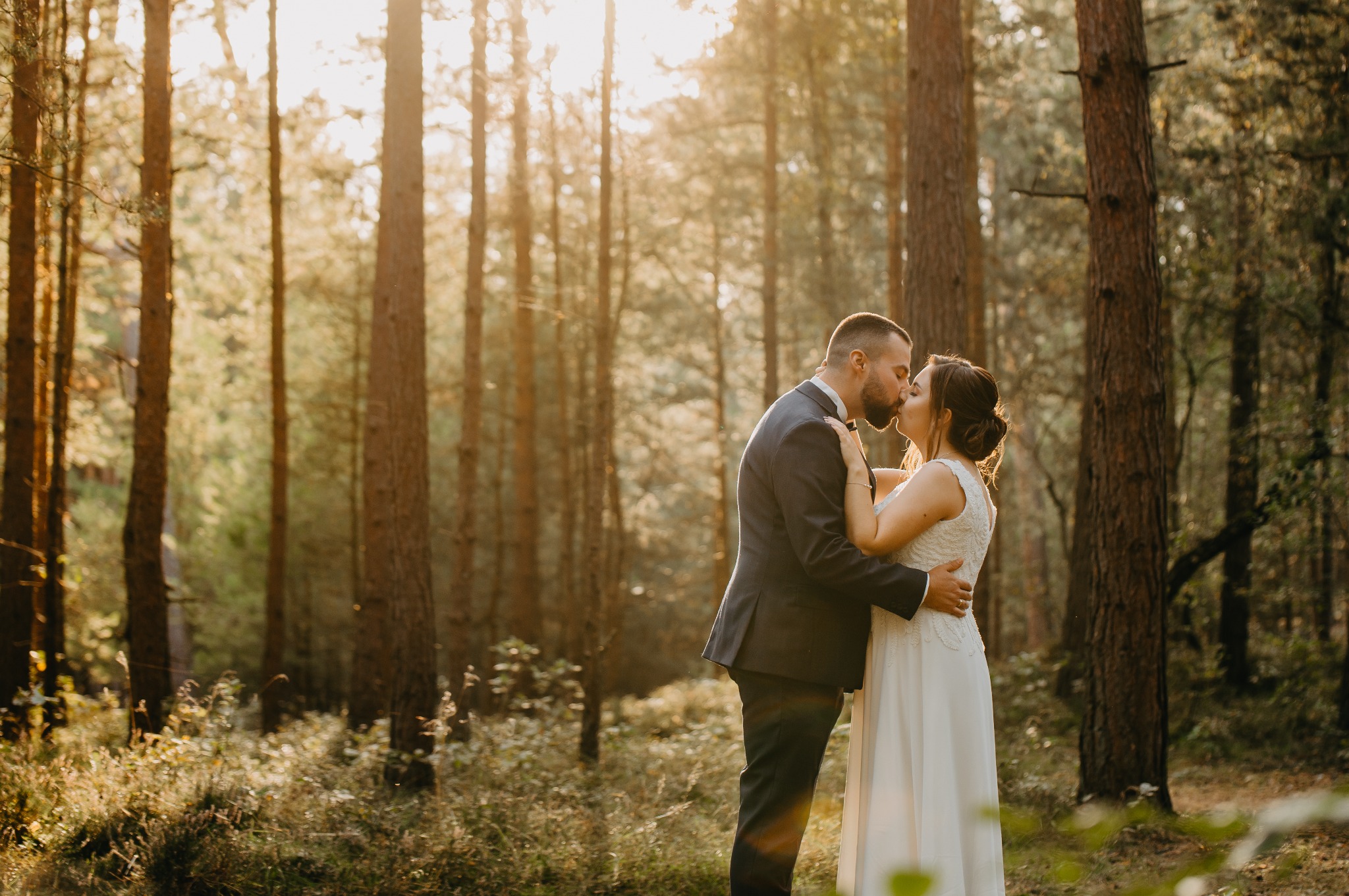 Pocałunek nowożeńców w lesie iglastym oświetlonym promieniami zachodzącego słońca, panna młoda w białej sukni z koronkową górą i upiętymi włosami, pan młody w ciemnoszarym garniturze.