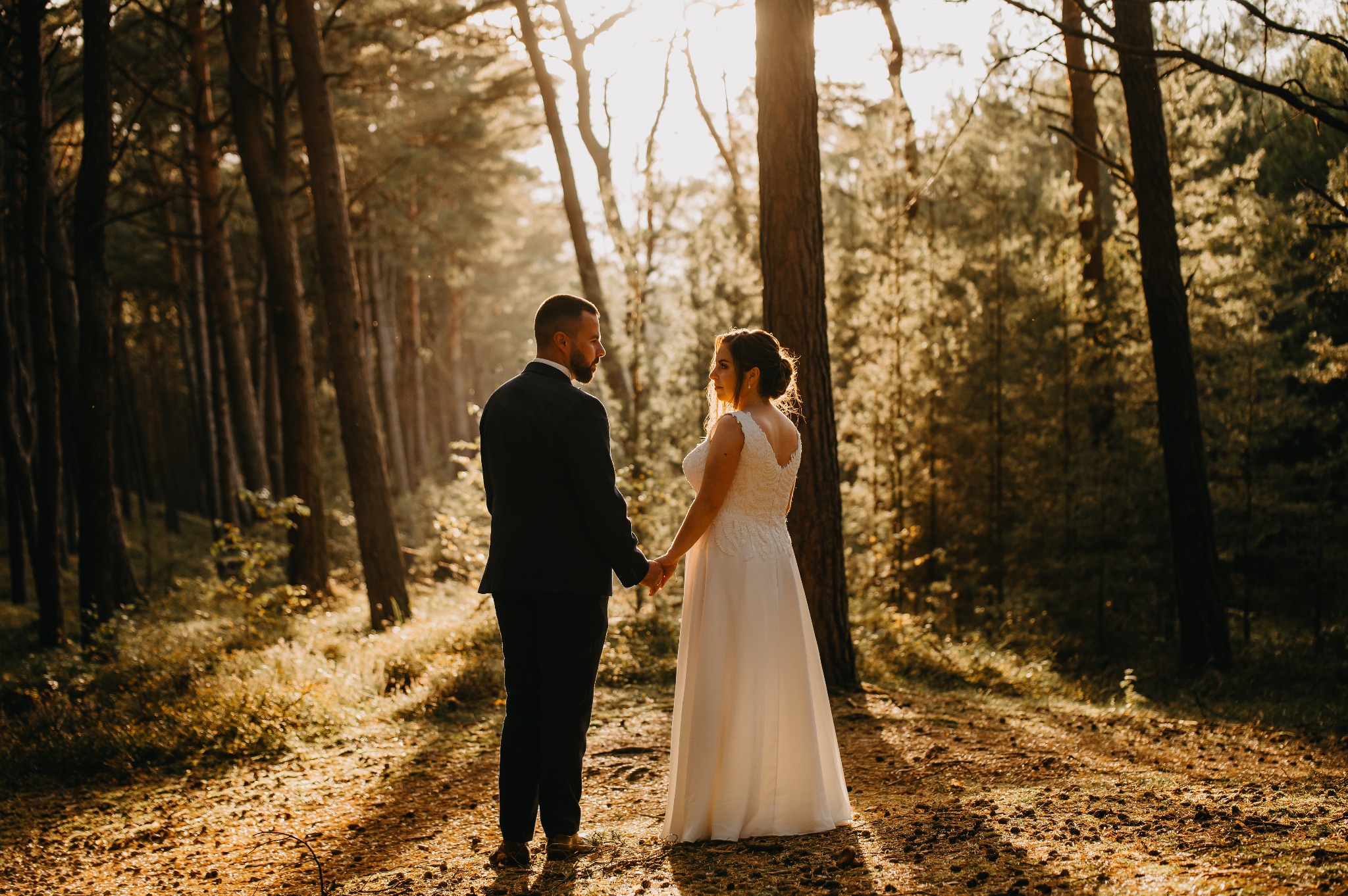 Para młoda trzymająca się za ręce w lesie, światło słoneczne przebija przez drzewa, ona w białej sukni z koronkowymi detalami na plecach, on w czarnym garniturze.