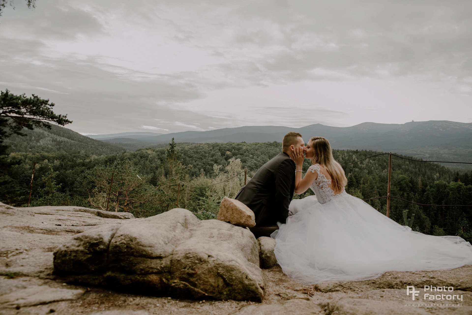 Nowożeńcy całujący się na skalnym tarasie z widokiem na zalesione góry w tle, suknia panny młodej z koronkową górą i długim tiulowym dołem.