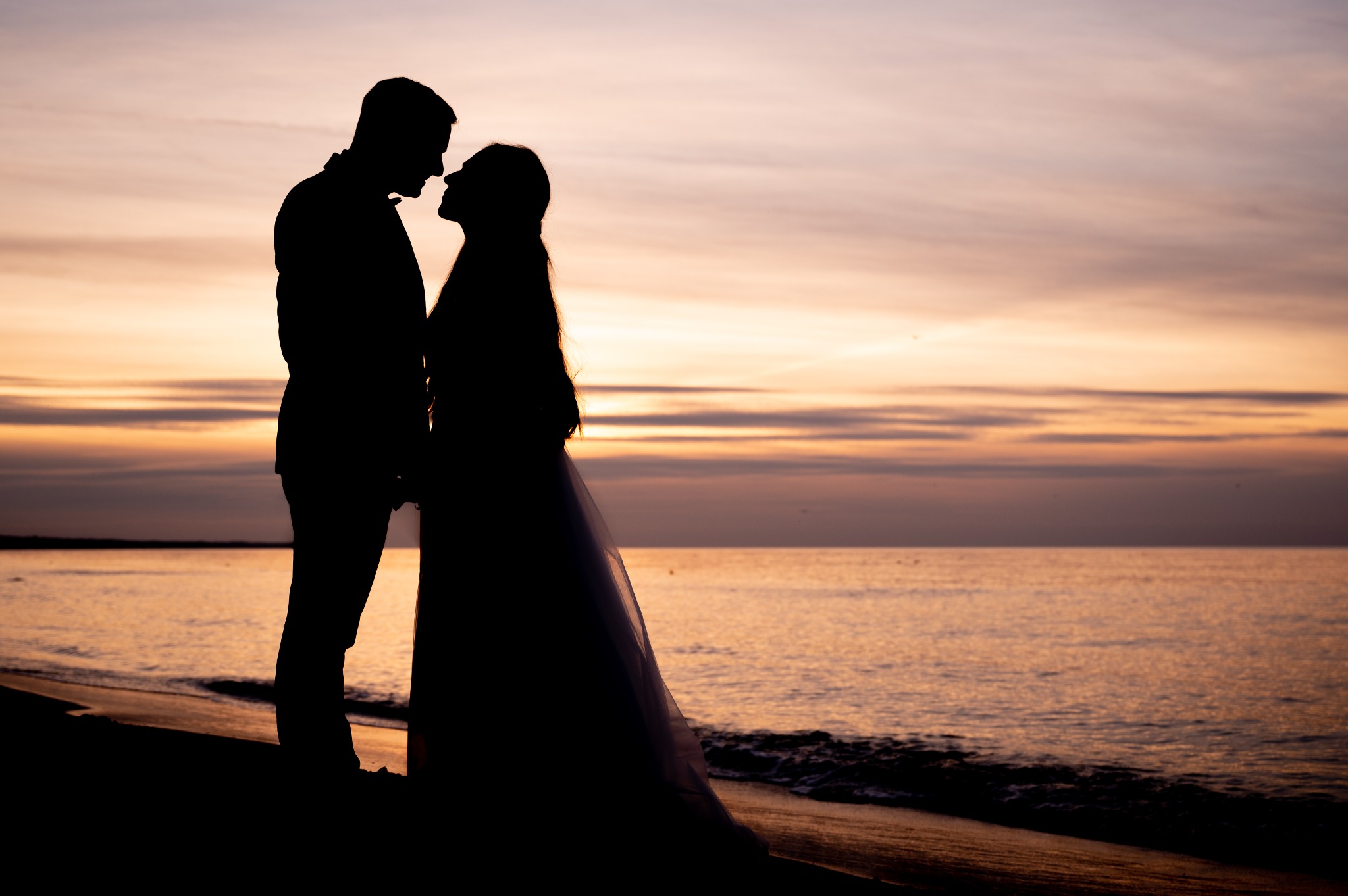 Sylwetki pary młodej na tle zachodzącego słońca nad morzem, panna młoda w długiej sukni, para stoi blisko siebie.