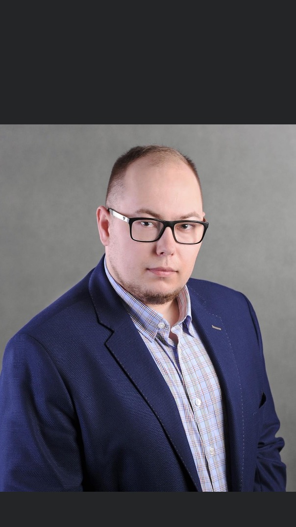 Portret mężczyzny w okularach i niebieskiej marynarce na szarym tle, z lekkim zarostem i poważnym wyrazem twarzy.