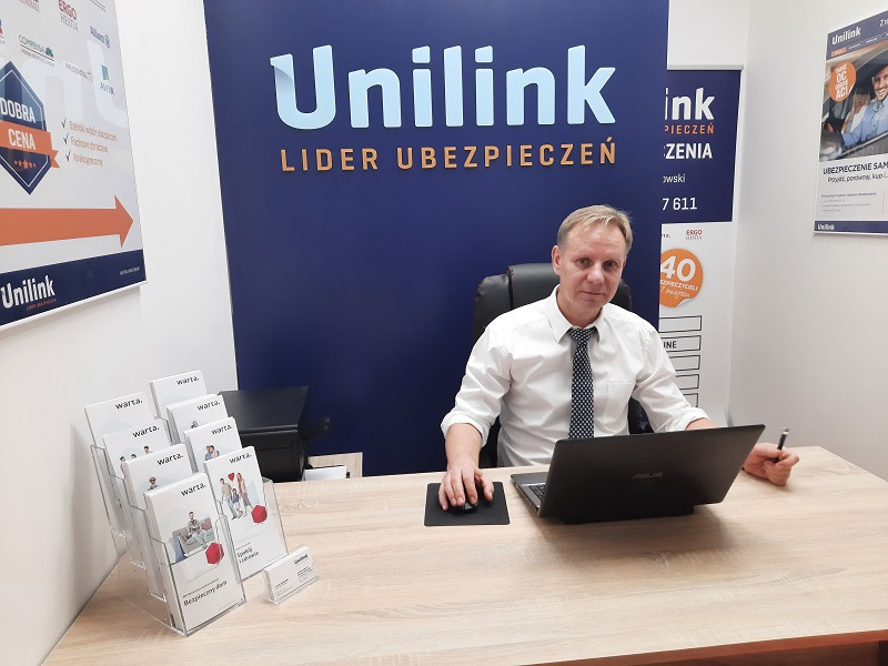 Agent Unilink siedzący za biurkiem z laptopem, w tle logo firmy i materiały informacyjne Warta.