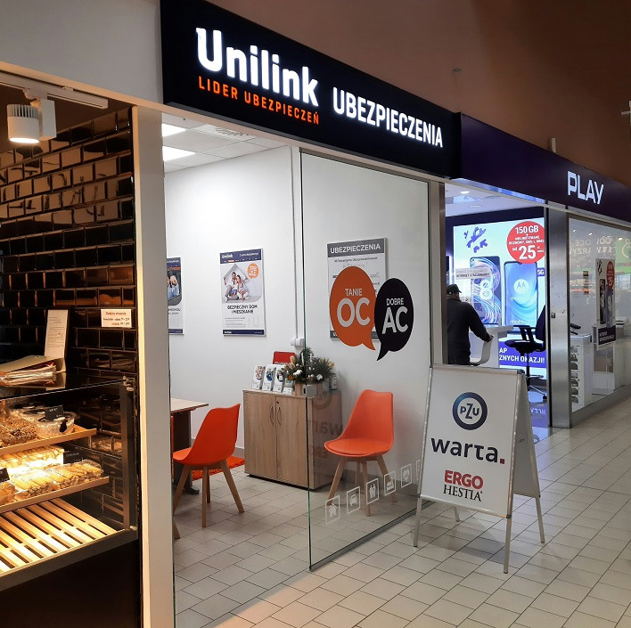Biuro ubezpieczeniowe Unilink w centrum handlowym, widoczne plakaty z ofertą OC/AC, stojak reklamowy z logo PZU, Warta i Ergo Hestia.
