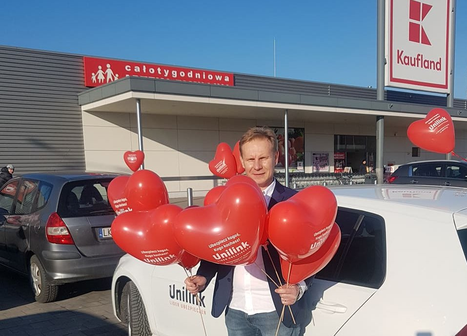 Mężczyzna w Kraśniku trzymający czerwone balony w kształcie serc z logo firmy ubezpieczeniowej Unilink, stojący przed samochodem z naklejką reklamową tej samej firmy na tle sklepu Kaufland.