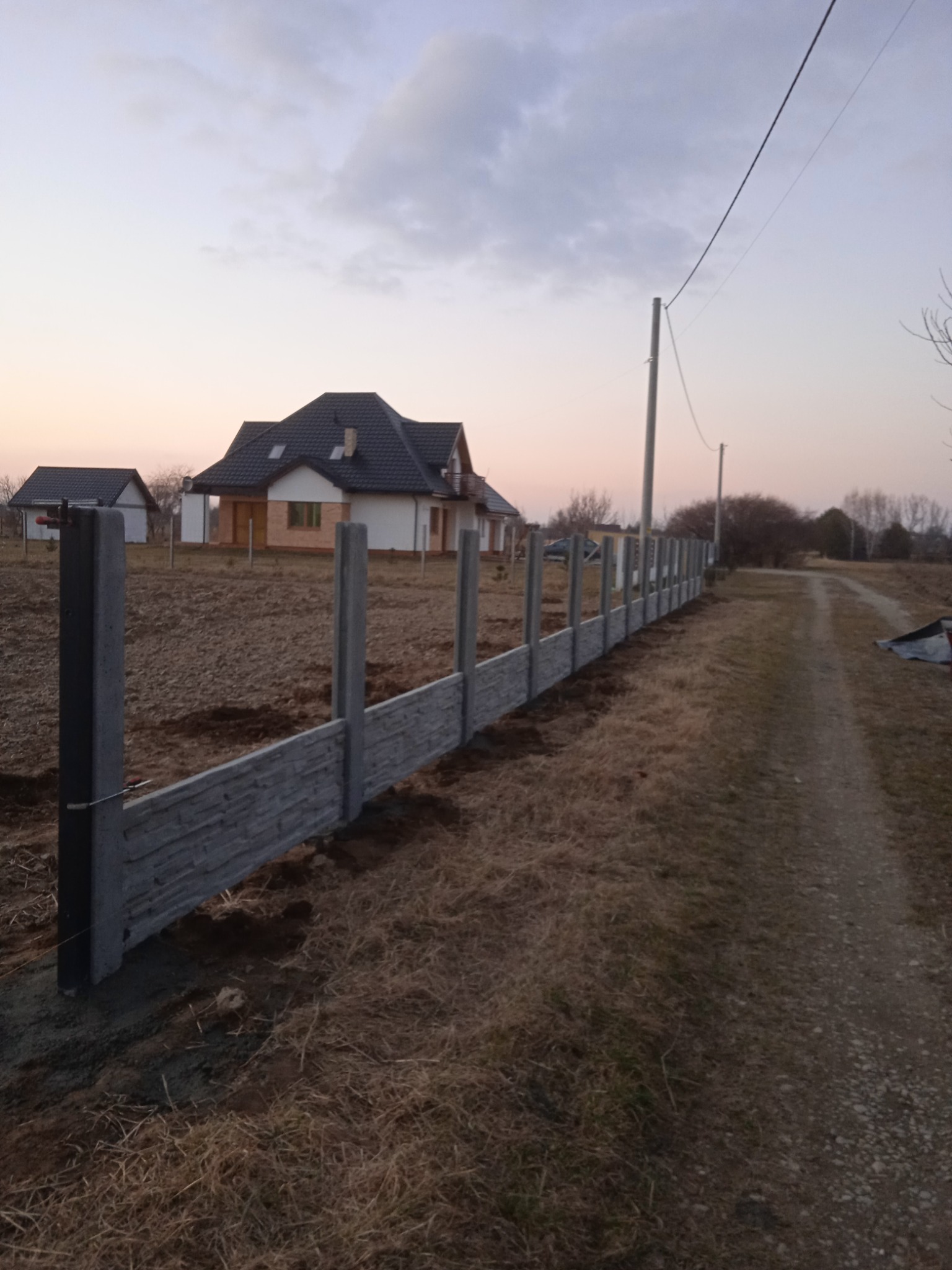 Fragment powstającego ogrodzenia z betonowych paneli imitujących kamień, widoczne słupki i częściowo zamontowane przęsła, w tle domy jednorodzinne i polna droga.