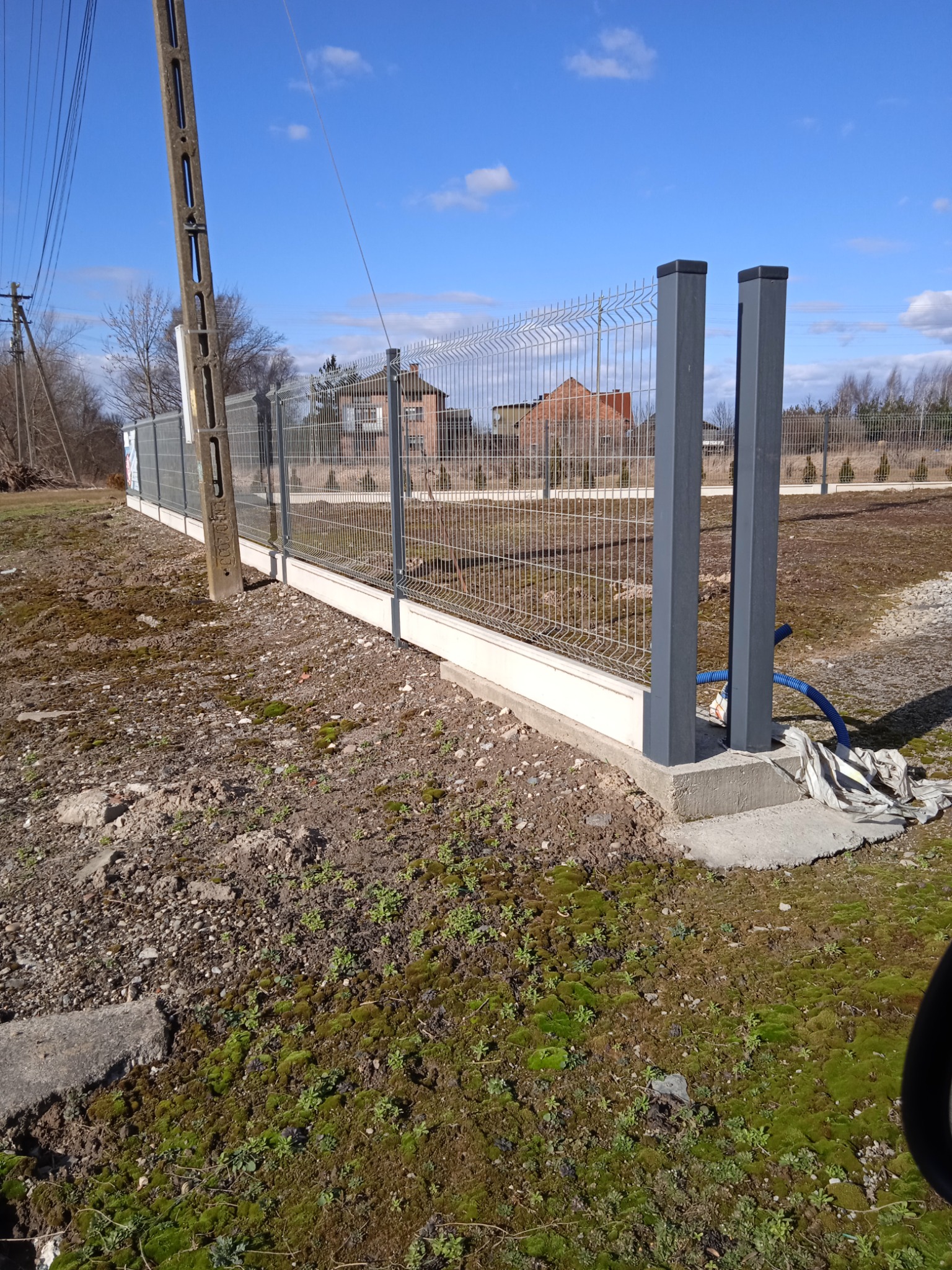 Nowoczesne ogrodzenie panelowe z szarą podmurówką i słupkami, widoczne na tle domu i błękitnego nieba, z fragmentem nieuporządkowanego terenu przed ogrodzeniem.