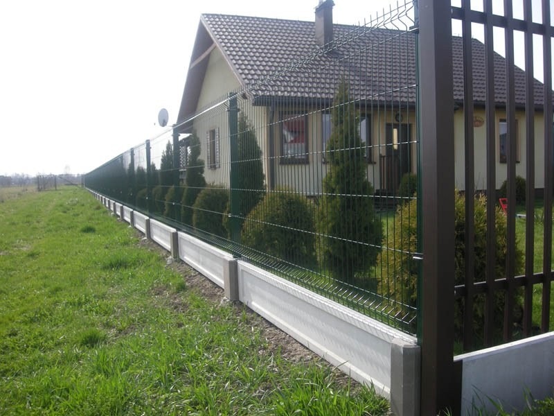 Długie, zielone ogrodzenie panelowe z podmurówką betonową, widok z zewnątrz posesji, w tle dom z brązowym dachem i zielonymi tujami.