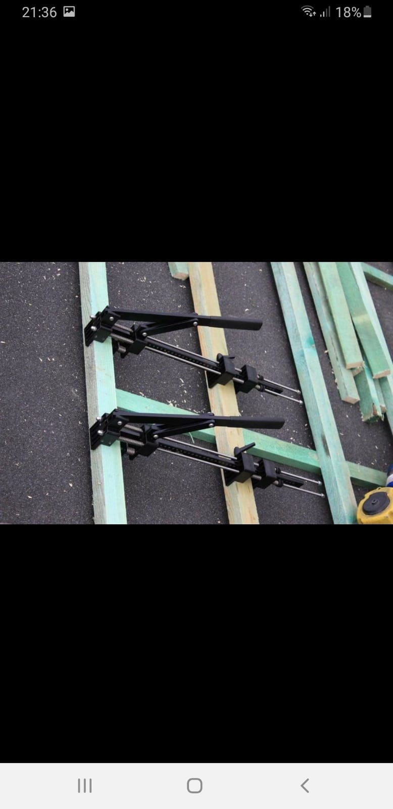 Dwa czarne zaciski dekarskie mocują drewniane łaty do dachu pokrytego papą, obok leżą dodatkowe łaty pomalowane na zielono.